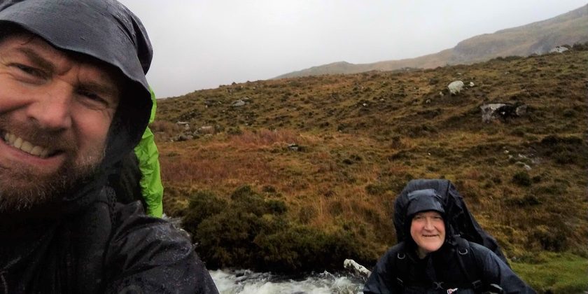 Man taking a selfie in rain gear and woman smiles on as they take part in a walking challenge
